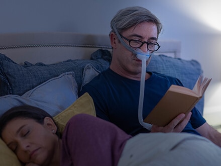 El paciente se beneficia de la terapia mientras lee un libro en la cama utilizando la mascarilla nasal minimalista ResMed AirFit N30