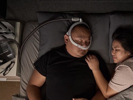 La pareja duerme en una cama. El marido utiliza una mascarilla de almohada nasal ResMed AirFit P30i como terapia.