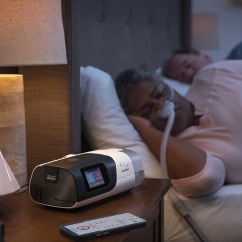 Un dispositivo de CPAP y un teléfono móvil sobre una mesilla de noche; al fondo, una mujer duerme junto a su pareja mientras lleva una mascarilla nasal de CPAP.