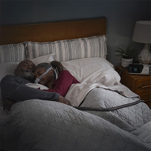 Una pareja se abraza mientras duerme en la cama, la mujer lleva una mascarilla CPAP.