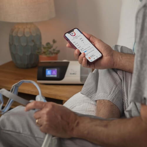 Una persona sentada en la cama con una mascarilla CPAP en una mano y un teléfono con una aplicación de terapia PAP en la otra.